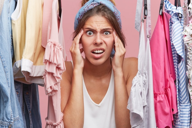 Mulher nervosa, mantendo as mãos no rosto, parecendo aterrorizada em pé perto do guarda-roupa com roupas, percebendo que não tem nada para vestir para se encontrar com os amigos. Conceito de emoções humanas negativas