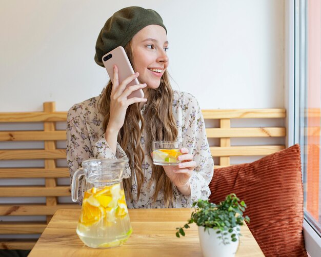 Mulher no meio de um tiro sentado à mesa segurando um copo de limonada e falando ao telefone