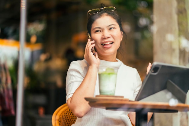 Mulher nômade digital asiática, atraente e inteligente, vestindo roupas casuais, trabalho remoto, novo estilo de vida normal, uso manual, tablet, teleconferência, reunião de videochamada ao vivo, remota no coffeeshop cafe