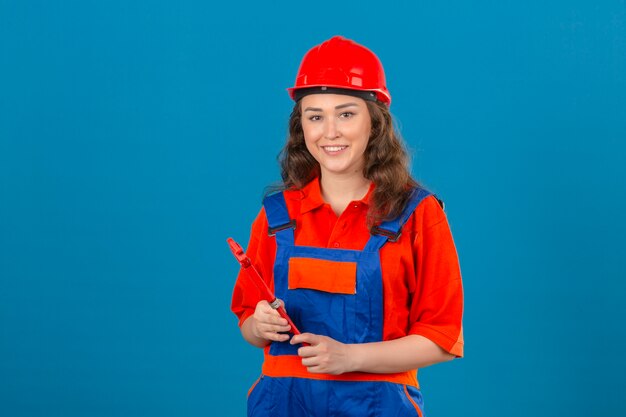 Mulher nova do construtor no uniforme da construção e no capacete de segurança que estão com a chave ajustável que olha feliz e sorrindo amigável sobre a parede azul isolada