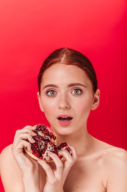 Mulher nua chocada segurando granada saborosa Foto de estúdio de menina adorável com romã expressando emoções surpresas em fundo vermelho