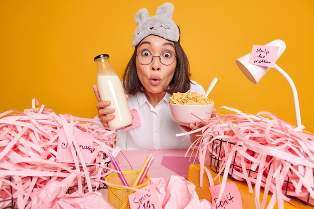 Mulher olha chocada com a câmera segura a tigela de cereais e a garrafa de leite se senta na área de trabalho com papel cortado rosa em torno de obras em home office isolado em amarelo.