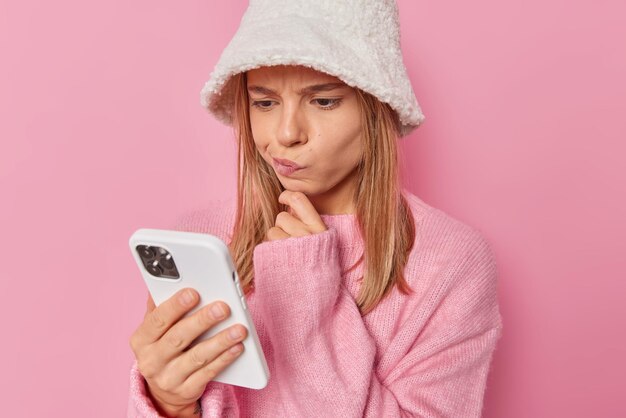 mulher olha para a tela do smartphone lê notificação recebe mensagem de pessoa desconhecida usa chapéu branco e o suéter isolado no rosa tem expressão duvidosa