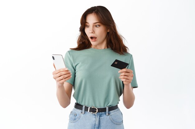 Mulher olhando chocada para a tela do smartphone enquanto paga com cartão de crédito, tendo problemas com conta bancária, transação de dinheiro, de pé contra uma parede branca