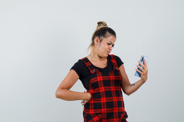 Mulher olhando para o celular com vestido de avental e parecendo hesitante, vista frontal.