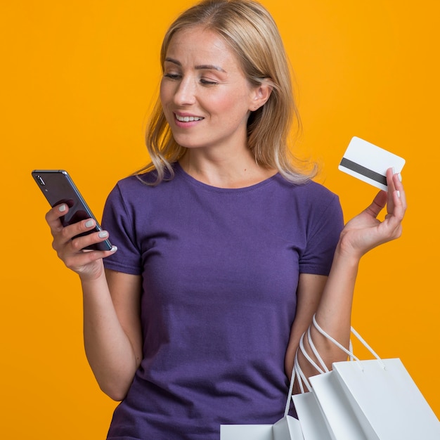 Mulher olhando para o smartphone, segurando um cartão de crédito e sacolas de compras