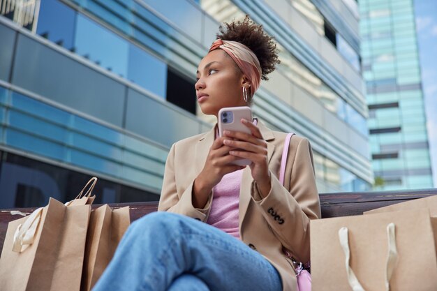 mulher olhando pensativamente para longe usa telefone celular para navegar em redes sociais poses no banco com muitas sacolas de papel em volta aproveitando o tempo de lazer