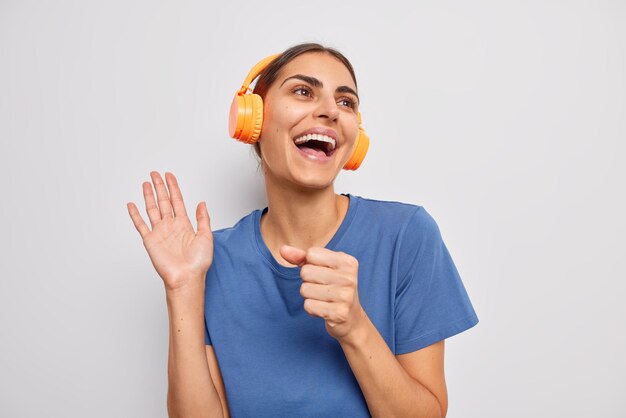 Mulher otimista e despreocupada mantém a palma da mão levantada perto da boca, como se o microfone cantasse a música favorita junto, vestida com uma camiseta azul casual isolada sobre o fundo branco, usando fones de ouvido laranja sem fio