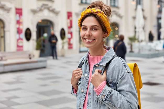 Mulher otimista positiva tem uma expressão feliz, usa roupas elegantes, tem uma pequena mochila amarela nas costas, perambula pelas ruas da cidade