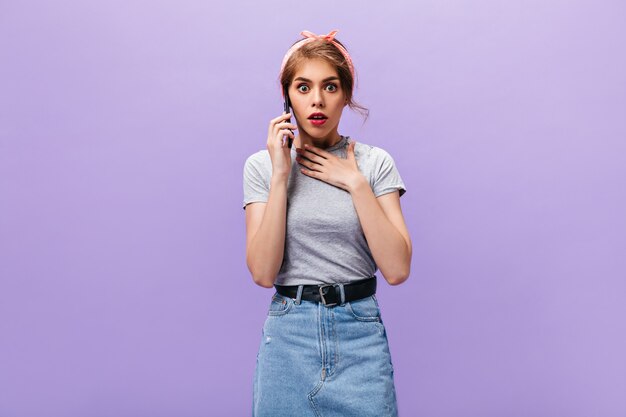 Mulher parece chocada enquanto fala ao telefone. Menina elegante com bandana rosa, camiseta cinza e saia jeans clara posing.n com fundo roxo.