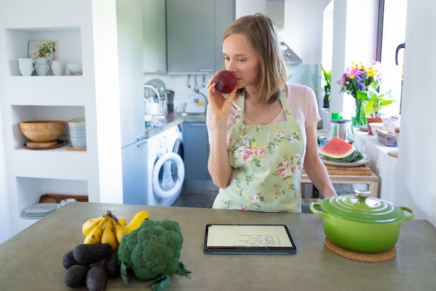 Mulher pensativa cheirando frutas enquanto cozinha na cozinha, usando o tablet perto da panela e legumes frescos no balcão. Vista frontal. Cozinhando em casa e conceito de alimentação saudável