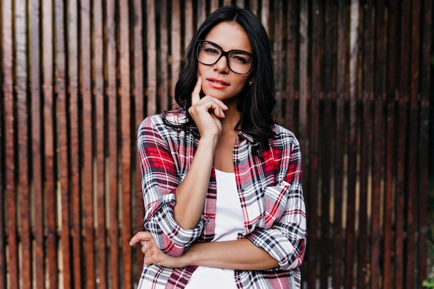 Mulher pensativa de camisa xadrez, tocando sua bochecha. Tiro ao ar livre de garota latina posando com expressão de rosto suspeito.