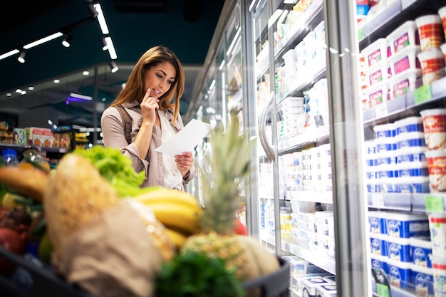 Mulher pensativa no supermercado segurando uma lista e lendo os itens de compras que está prestes a comprar