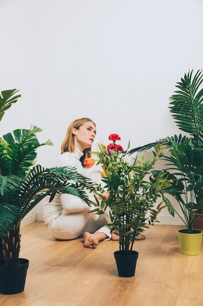 Mulher pensativa, sentada no chão com flores perto de plantas verdes
