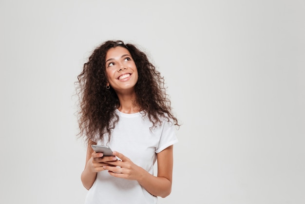 Mulher pensativa sorridente segurando o smartphone nas mãos e olhando sobre fundo cinza