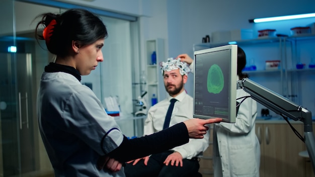 Mulher pesquisadora olhando para monitor analisando tomografia cerebral enquanto uma colega de trabalho discute com a paciente sobre os efeitos colaterais