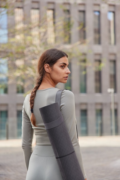 Foto grátis mulher posa com colchonete de borracha vai para a academia se prepara para o treino vestida com roupas esportivas caminha no centro da cidade