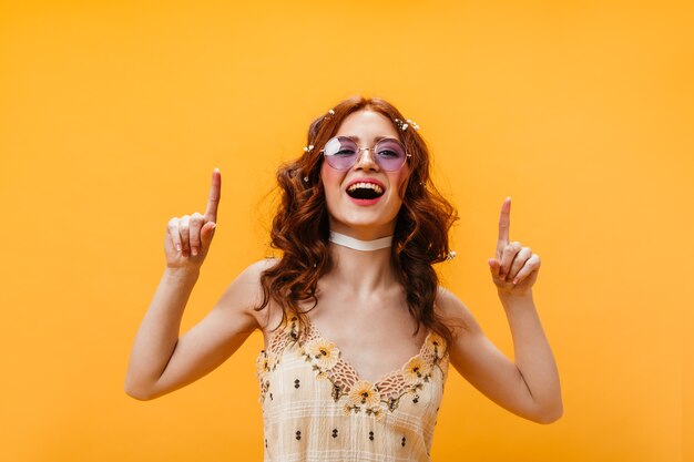 Mulher positiva aponta os dedos para cima. mulher de óculos lilás e top amarelo, posando em fundo laranja.