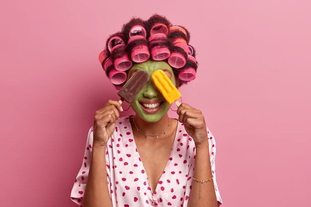 Mulher positiva cobre os olhos com dois deliciosos sorvetes, sorri feliz, aplica máscara facial verde e rolos de cabelo, vestida com manto casual, posa