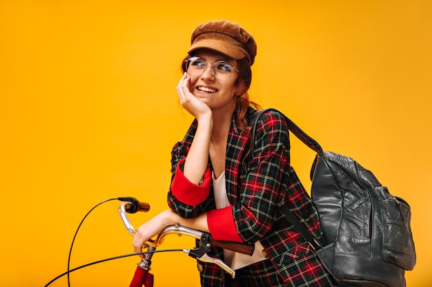 Mulher positiva com boné de veludo e óculos posando sonhadoramente com uma bicicleta