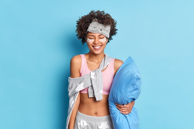 Foto grátis mulher positiva com cabelo encaracolado acorda de manhã entusiasmada e satisfeita após uma boa noite de sono em poses de pijama segurando travesseiro isolado sobre a parede azul