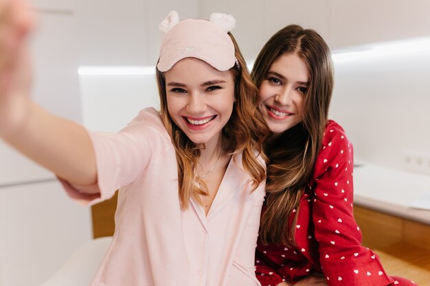 Mulher positiva de cabelos escuros posando enquanto a irmã fazendo selfie. Jovens sonhadoras sorrindo de manhã cedo.