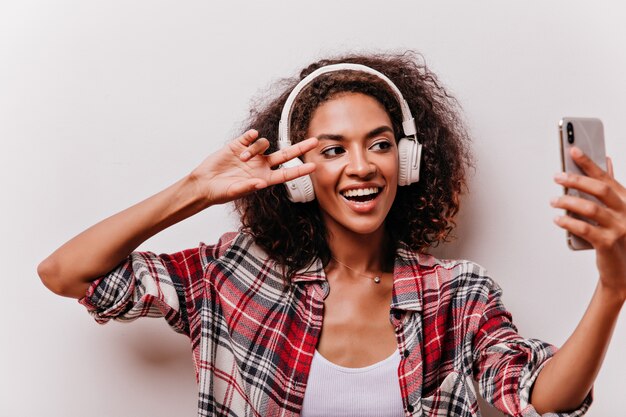 Mulher positiva de olhos pretos fazendo selfie com o símbolo da paz e rindo. Jocund menina encaracolada em grandes fones de ouvido se divertindo.