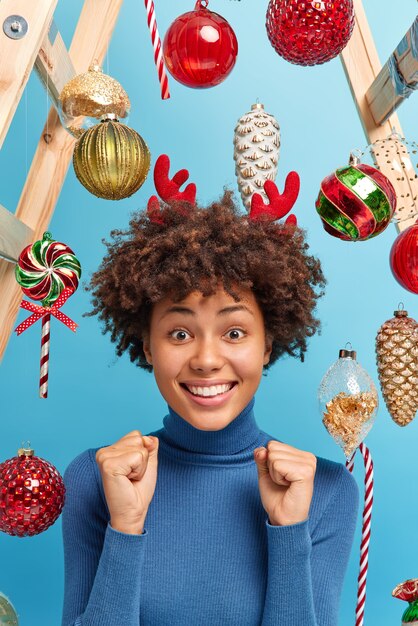 Mulher positiva de pele escura com cabelo encaracolado em gola rolê casual fecha os punhos antecipa para o milagre se prepara para as férias de Natal vestida casualmente posa dentro de casa sobre os brinquedos de ano novo. Evento festivo.
