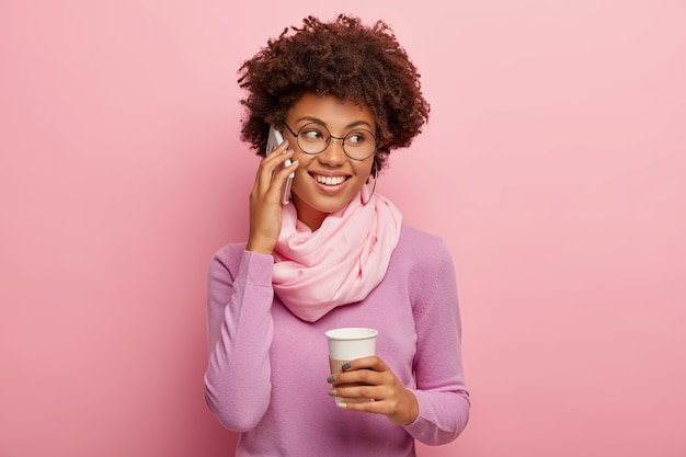 Mulher positiva de pele escura com corte de cabelo afro, fala pelo celular, segura café para viagem, gosta de uma conversa agradável, usa blusão casual e lenço de seda, sorri amplamente, discute algo