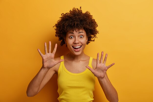 Mulher positiva de pele escura levanta as palmas das mãos, sente-se alegre, tem humor brincalhão, olha com expressão engraçada, usa camisa casual, isolada sobre parede amarela. Conceito de pessoas, emoções e felicidade