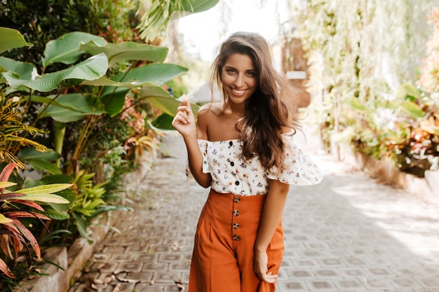 Mulher positiva em shorts largos laranja e blusa branca sorri coquete, segurando uma pequena flor nas mãos