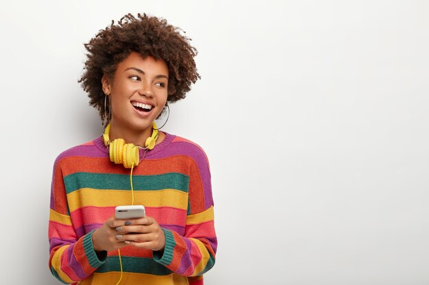 Mulher positiva ri com alegria, usa celular atualizado para bater papo online, vira as costas, usa fone de ouvido e suéter, isolado no branco