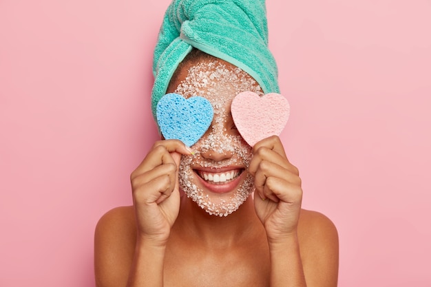 Foto grátis mulher positiva se diverte durante os tratamentos de beleza, mantém duas esponjas em forma de coração nos olhos, tem um sorriso largo, mostra os dentes brancos, toalha enrolada na cabeça, posa dentro de casa com o corpo nu