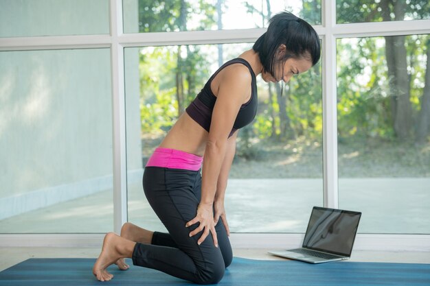 Mulher praticando ioga, fazendo exercícios de bloqueio abdominal para cima, uddiyana Bandha pose, malhando, vestindo roupas esportivas, assistindo a um vídeo tutorial de fitness online no laptop, fazendo exercícios em casa sentado.