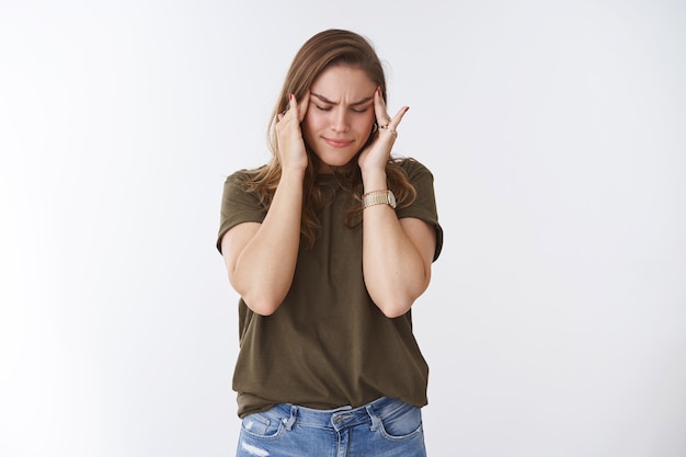 Mulher que sofre de uma enorme enxaqueca, sensação dolorosa, tocando as têmporas, não consegue conter a dor, fecha os olhos franzindo a testa intensamente sem foco, os analgésicos de forte dor de cabeça não funcionam, em pé, fundo branco doente