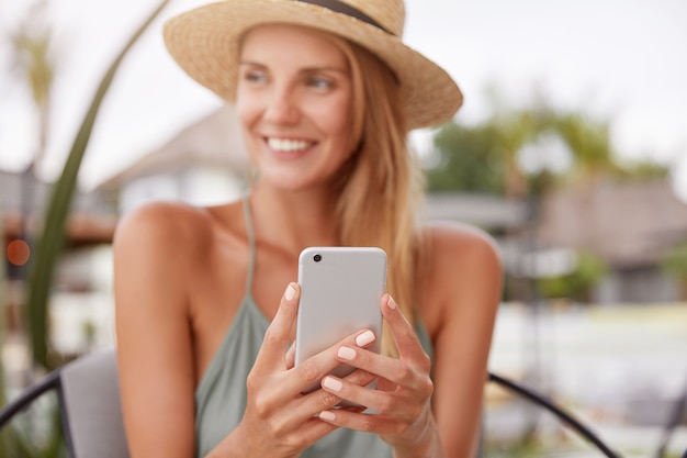 Mulher relaxada alegre usa telefone inteligente para bate-papo com amigos, senta-se em um café moderno ou um café com terraço. Linda mulher lê boas notícias no site da internet, aproveita o descanso de verão. Foco no celular