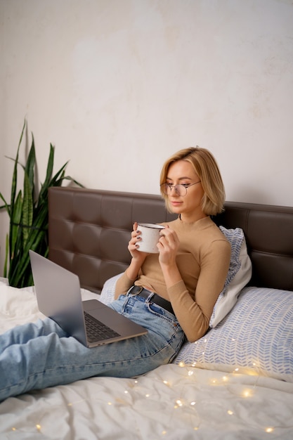 Mulher relaxando e bebendo uma xícara de café ou chá quente usando o computador portátil no quarto.