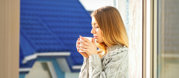 Mulher relaxando na varanda com café. uma bela jovem com um suéter segura uma xícara de café em pé perto da janela pela manhã.
