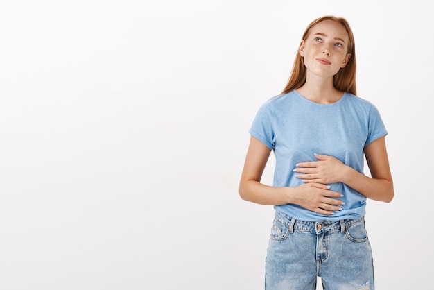 Foto grátis mulher relembrando comida saborosa que comia em restaurante novo esfregando barriga ou barriga olhando para canto superior esquerdo nostálgica e feliz gostando de comer deliciosas refeições em pé de camiseta azul