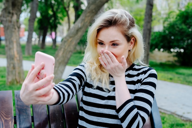 Mulher rindo tomando selfie com smartphone enquanto está sentado ao ar livre em um banco no topo listrado