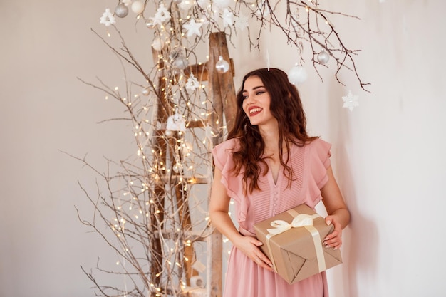Mulher romântica em um vestido rosa com caixa de presente de natal está posando ao lado de decorações festivas, parede branca.