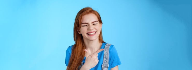 Foto grátis mulher ruiva alegre e despreocupada, sorriso sincero, apontando para a esquerda, introduza o espaço da cópia com alegria, feche os olhos