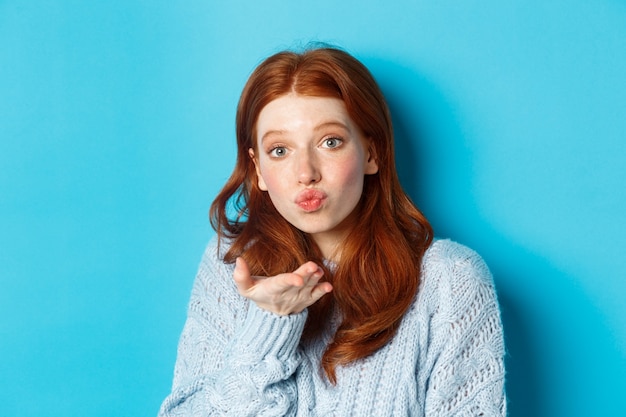 Mulher ruiva boba de suéter, mandando beijo no ar para a câmera com lábios franzidos, de pé contra um fundo azul