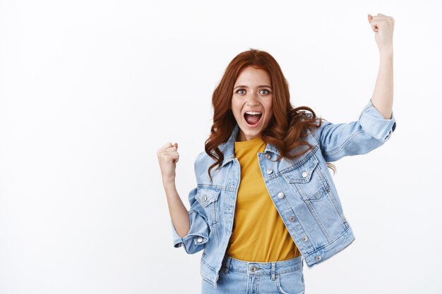 Mulher ruiva bonita animada em jaqueta jeans, levante as mãos em sim, gesto de viva, grito de soco encorajado, torcendo pelo time favorito, sorrindo satisfeito, alcance o objetivo, comemore o sucesso