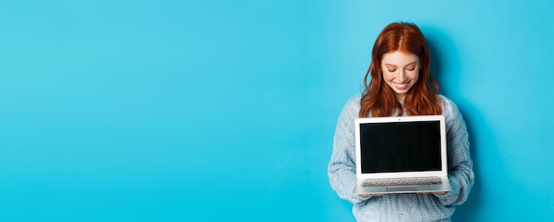 Mulher ruiva bonita de suéter mostrando e olhando para a tela do laptop com um sorriso satisfeito demonstrando