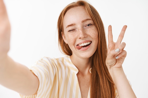 Mulher ruiva bonita e elegante com sardas fofas, mostrando a língua e sorrindo de alegria, piscando alegremente, mostrando o sinal da paz, tirando selfie no smartphone