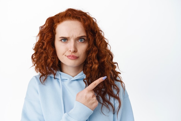 Mulher ruiva com a testa franzida e decepcionada apontando o dedo para algo ruim, repreendendo ou reclamando, parece chateada e insatisfeita, parede branca