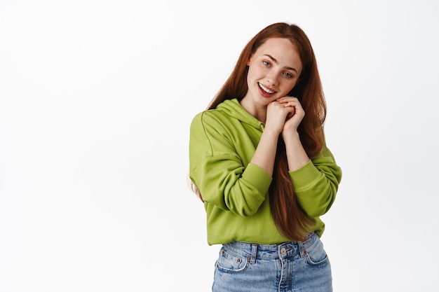 Mulher ruiva concurso olhando e admirando algo, sorrindo romântico e adorável, de pé em roupas casuais contra fundo branco.