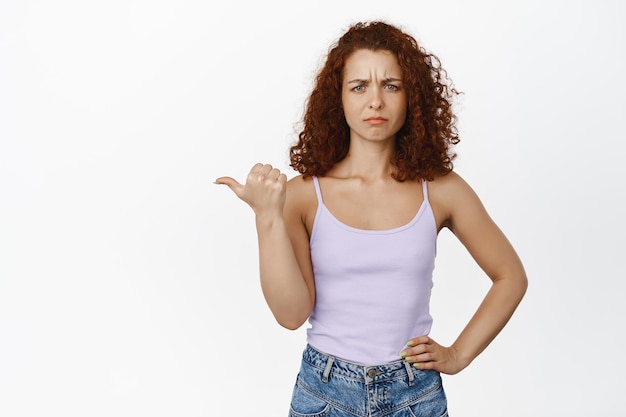 Mulher ruiva confusa e descontente franzindo a testa chateada, apontando para a esquerda com cara de raiva questionada, não gosta de smth, de pé contra um fundo branco.