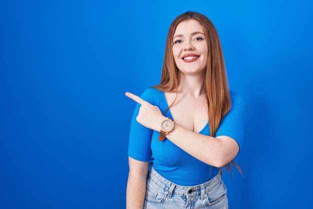 Mulher ruiva em pé sobre fundo azul alegre com um sorriso no rosto apontando com a mão e o dedo para o lado com expressão feliz e natural